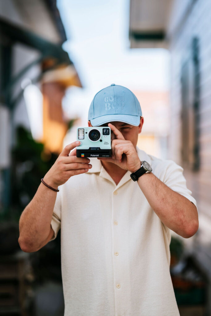 Homme en train de prendre une photo avec un polaroide. Il porte une casquette bleu ciel My CAP avec brodé "Good Boy" dessus.